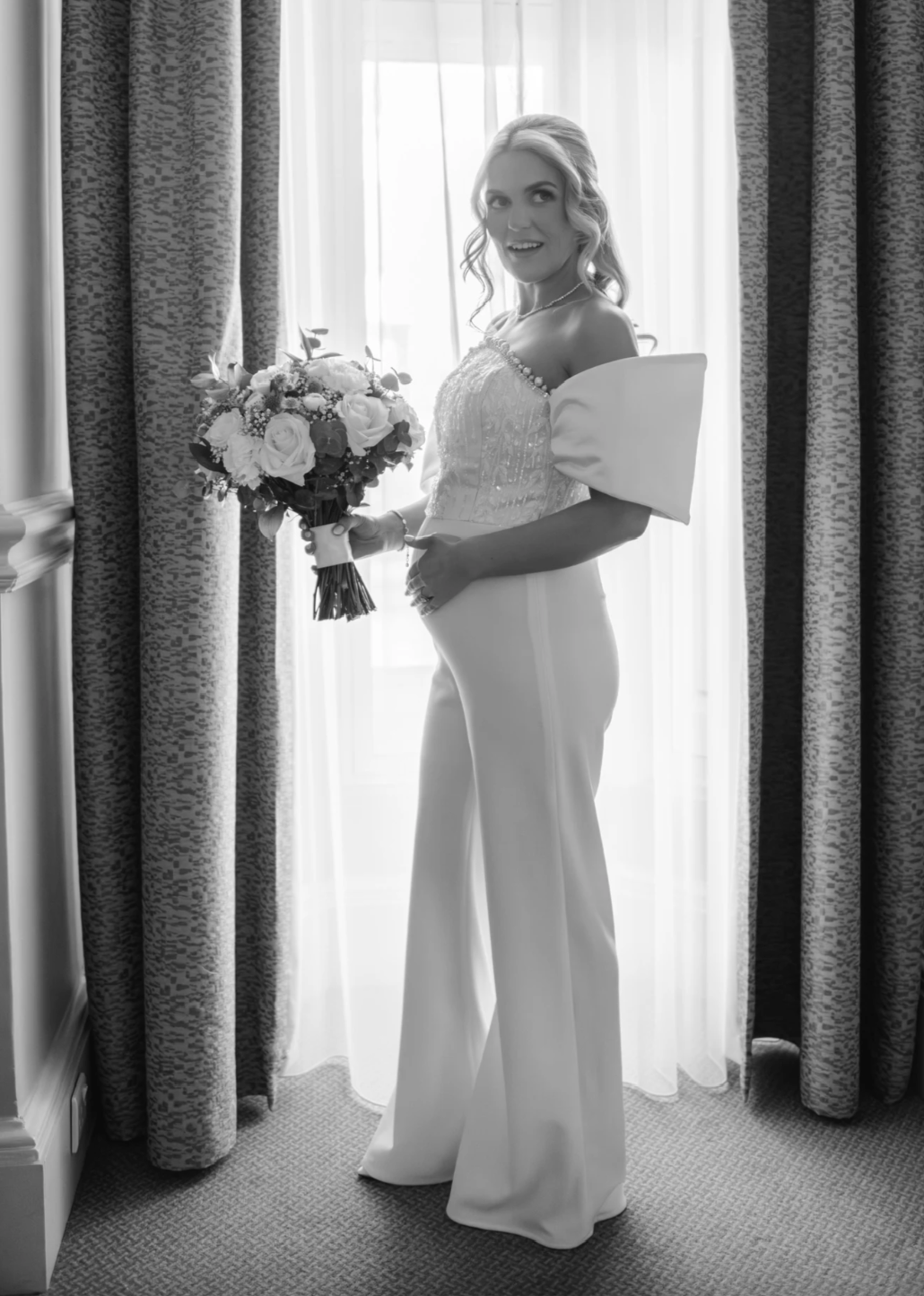 Nicole Fletcher wears a bespoke bridal maternity jumpsuit by Rebecca Tembo. The jumpsuit is off-shoulder, strapless with a beaded bodice and crepe pants. The colour is ivory, the photo is in black and white.