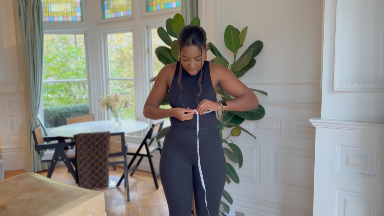 Load video: Rebecca is wearing a black outfit in a living room, holding a measuring tape and showing viewers how to take their measurements